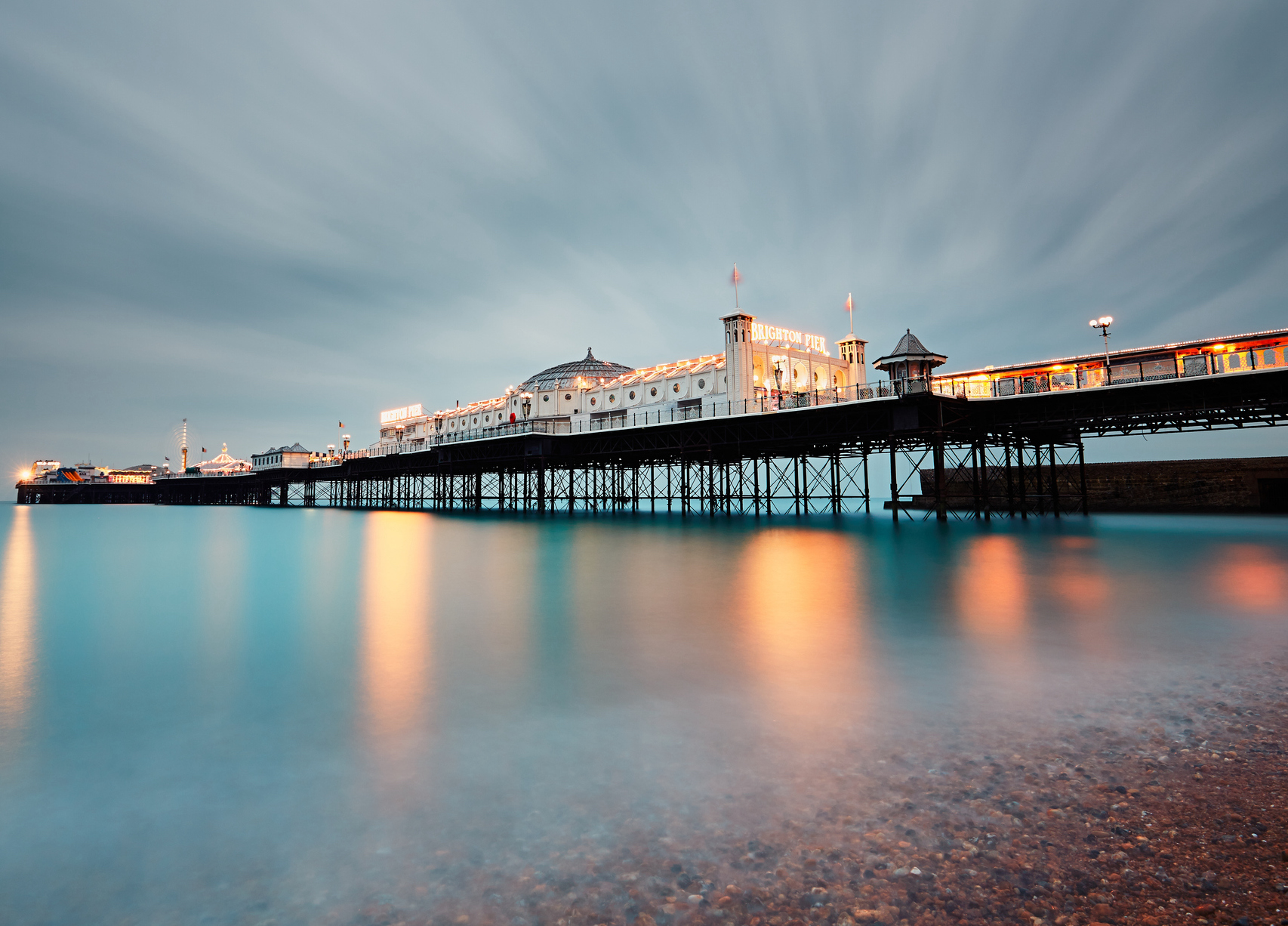 Brighton Pier lit up at dusk, with lights reflected in the body of still water below the pier